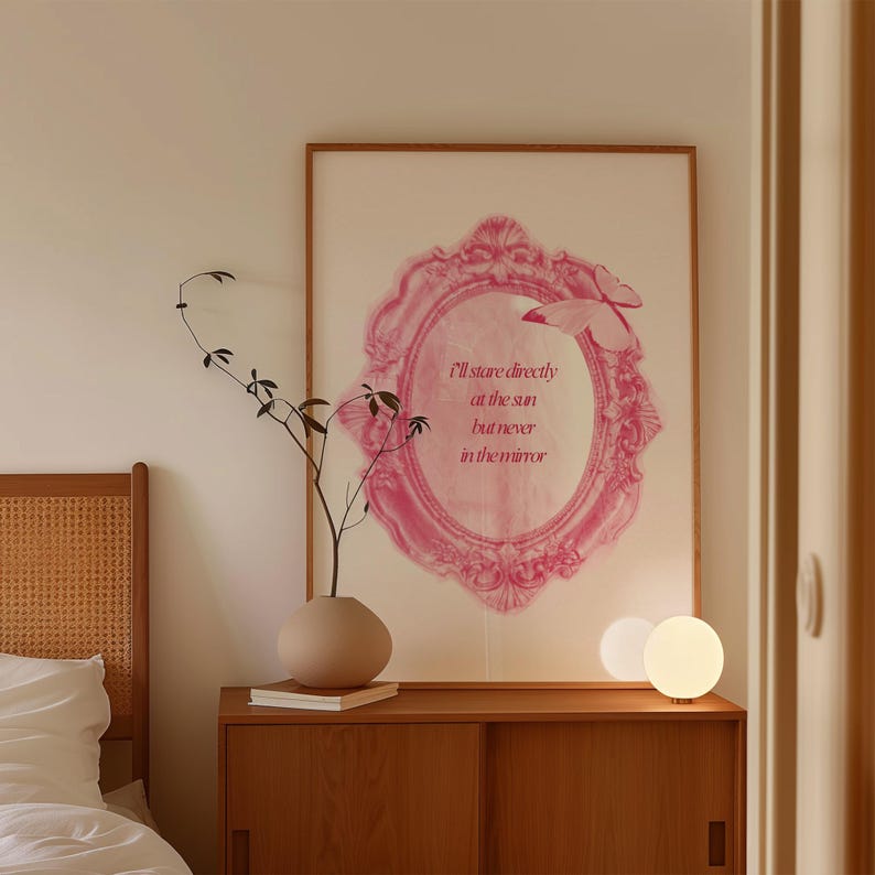 Pode incluir: Um interior de quarto com uma mesa de cabeceira de madeira, uma l&acirc;mpada branca, um vaso com um galho seco e uma impress&atilde;o emoldurada com uma moldura rosa adornada e o texto "Vou olhar diretamente para o sol, mas nunca para o espelho".