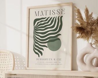 May include: A framed art print with a green abstract design and the text "MATISSE" and "BERGGREEN & CIE" in a minimalist style. The print is displayed on a light wood shelf with a vase of dried pampas grass.