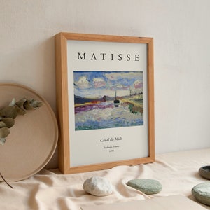 Pode incluir: Uma impressão emoldurada de uma pintura de Henri Matisse intitulada "Canal du Midi". A pintura representa um canal com um barco e um céu nublado. A moldura é de madeira e tem um acabamento natural.