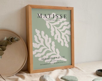 May include: A framed print of a Matisse artwork titled "Papiers Decoupés" with a green background and white abstract shapes. The print is framed in a light wood frame and includes the text "BERGGRUEN & CIE" and the dates "Du Vendredi 22. Février au Samedi 24 Mars 1973".