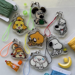 May include: A collection of seven clear acrylic keychains featuring various cartoon characters, including a yellow dog, a white dog with black ears, a yellow creature eating, and a white puppy, each attached to a colourful nylon cord loop.