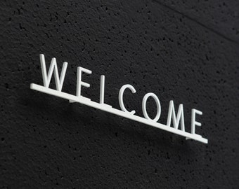 Könnte beinhalten: Ein weißes Metallschild mit dem Wort "WELCOME" an einer schwarzen, strukturierten Wand.
