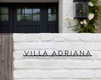 May include: A white brick wall with a black metal sign that reads "VILLA ADRIANA".