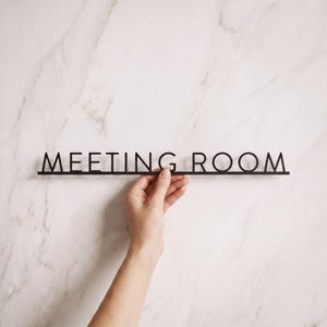 May include: A black, thin, linear sign featuring the words MEETING ROOM in a clean, sans-serif capital font. The letters are connected by a single horizontal bar at the base, creating a unified, minimalist piece of signage for office spaces.