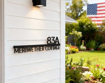 May include: Black metal house number sign with the address "83A MERIWETHER COTTAGE" mounted on a white clapboard wall. A black square light fixture is above the sign. An American flag waves in the background.