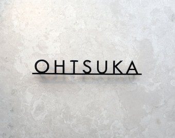 Puede incluir: Letrero de metal negro con el nombre "OHTSUKA" en una fuente minimalista. Las letras están conectadas por una línea fina y recta. El letrero está montado en una pared gris claro texturizada.