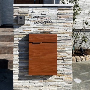 May include: A wooden mailbox with a modern design is mounted on a stone wall. The wall is made of stacked stone tiles in various shades of gray and brown. A metal nameplate with the word "Tomioka" is mounted above the mailbox.