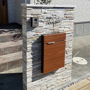 May include: A stone wall with a wooden mailbox and a doorbell. The wall is made of stacked stone tiles in shades of gray and brown. The mailbox is a rectangular wooden box with a metal handle. The doorbell is a silver rectangle with a small camera lens.