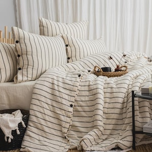 May include: A bed with a cream-colored duvet and pillows, all with black horizontal stripes. The pillows have black buttons. A woven tray with a black cup and small vase sits on the duvet. A black basket with a white towel is in the foreground.