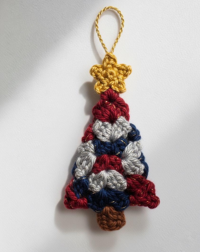 May include: A handmade crochet Christmas tree ornament. The tree is made of red, white, and blue yarn, with a gold star on top and a brown trunk. A gold string is attached for hanging.