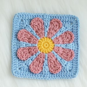Può includere: Un pezzo quadrato all'uncinetto con un motivo a margherita. Il centro del fiore è giallo, circondato da petali rosa. Lo sfondo è azzurro, misura circa 15 cm quadrati.