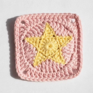 May include: A square, crocheted item featuring a yellow star design centered on a pink background. The handmade piece has a textured appearance with visible stitch details. The corners have small holes.
