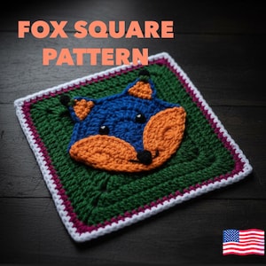Könnte beinhalten: Ein gehäkeltes Quadrat mit einem Fuchsmotiv. Der Fuchs hat einen blauen Kopf, orange Ohren und Schnauze sowie schwarze Augen und Nase. Das Quadrat ist grün mit einem weißen, rosa und weißen Rand. Der Text "FOX SQUARE PATTERN" steht oben.