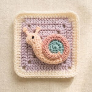 May include: A crocheted square featuring a snail design. The snail is in shades of pink, peach, and teal, set against a lavender and cream-colored background. The square is bordered with a cream-colored crochet stitch.