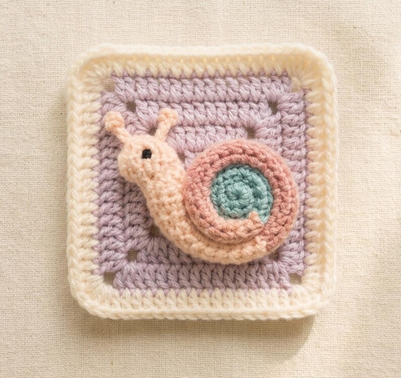 May include: A crocheted square featuring a snail design. The snail is in shades of pink, peach, and teal, set against a lavender and cream-colored background. The square is bordered with a cream-colored crochet stitch.