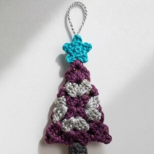 May include: A handmade crochet Christmas tree ornament. The tree is purple, gray, and gold, topped with a turquoise star and a silver hanging loop. The tree is approximately 4 inches tall.