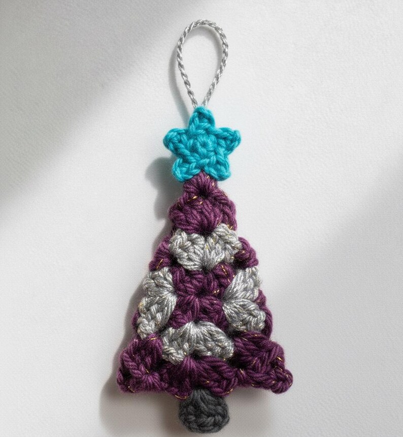 May include: A handmade crochet Christmas tree ornament. The tree is purple, gray, and gold, topped with a turquoise star and a silver hanging loop. The tree is approximately 4 inches tall.