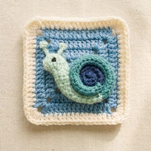 May include: A crocheted square featuring a snail design. The snail is light green with a dark blue shell, set against a light blue background. The square is bordered with a cream-colored crochet stitch.