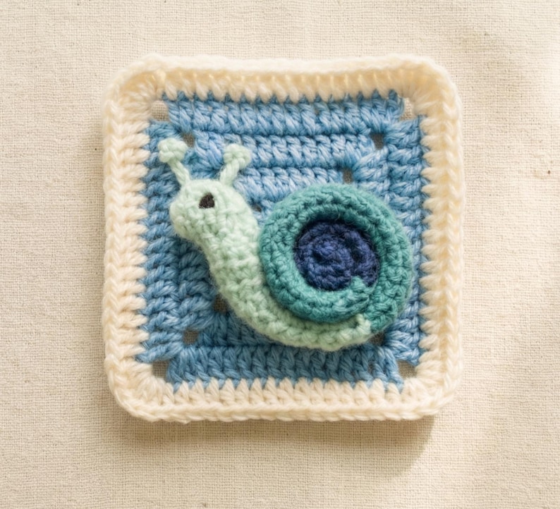 May include: A crocheted square featuring a snail design. The snail is light green with a dark blue shell, set against a light blue background. The square is bordered with a cream-colored crochet stitch.