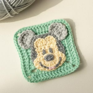 Könnte beinhalten: Ein quadratisches, gehäkeltes Applikationsmuster mit dem Gesicht von Mickey Mouse. Das Design verwendet hellgelbes, graues und rosa Garn für das Gesicht, mit einem grünen Rand. Eine Kugel aus grauem Garn befindet sich in der oberen linken Ecke.