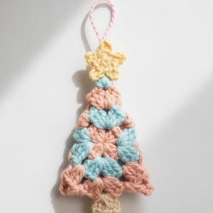 May include: A handmade crochet Christmas tree ornament. The tree is made of pink and blue yarn, topped with a yellow star and a pink hanging loop. The base of the tree is beige. The ornament is designed to be hung.