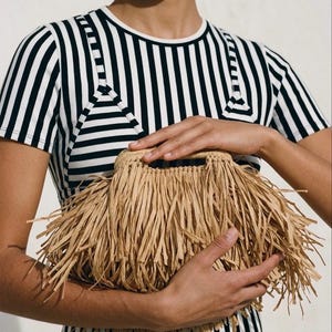 May include: A natural raffia handbag with a fringe design. The bag is held by a person wearing a black and white striped top. The bag has a woven handle and a rectangular shape, with long raffia strands creating a fringe effect.