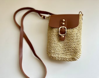 May include: A small straw crossbody bag with a brown leather flap and a gold buckle. The bag has a long brown strap.