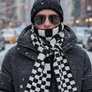 May include: A person wearing a black knitted beanie, aviator sunglasses, and a black and white chequered scarf. The person is also wearing a dark grey jacket. Snow is falling in the background.