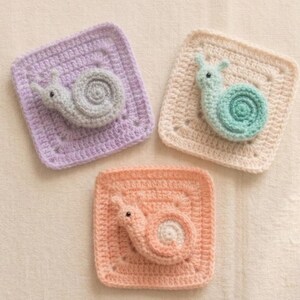 May include: Three handmade crochet squares, each featuring a snail design. The squares are in pastel colors: lavender, peach, and cream. The snails are gray, peach, and teal. The squares are approximately 4 inches by 4 inches.