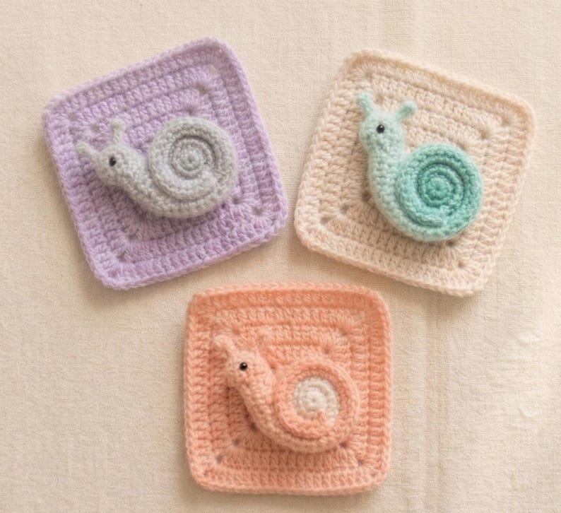 May include: Three handmade crochet squares, each featuring a snail design. The squares are in pastel colors: lavender, peach, and cream. The snails are gray, peach, and teal. The squares are approximately 4 inches by 4 inches.
