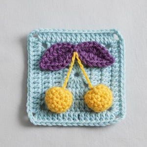 Peut inclure: Pièce carrée crochetée en bleu clair, ornée d'un motif de cerises jaunes avec des feuilles violettes. Les cerises sont rondes et texturées, avec des tiges jaunes. Les feuilles sont violettes foncées. La pièce est probablement faite à la main.