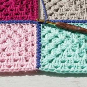 May include: Close-up of a colorful crochet project in progress. The work features squares in shades of pink, teal, gray, and fuchsia, joined with blue yarn. A wooden crochet hook is inserted into the teal section, with a strand of blue yarn attached.
