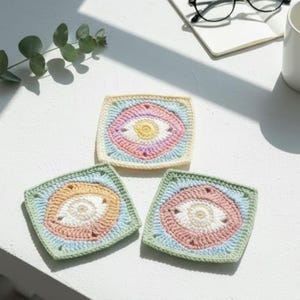 Könnte beinhalten: Drei quadratische Häkeluntersetzer in Pastellfarben: Gelb, Rosa, Blau und Grün. Jeder Untersetzer hat ein zentrales augenähnliches Design. Die handgefertigten Untersetzer sind auf einer weißen Oberfläche platziert, passend für die Heimdekoration.