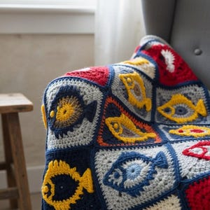 May include: A handmade crochet blanket featuring a repeating fish pattern. The blanket is made of squares in various colors, including navy blue, red, yellow, and gray. The fish are yellow and blue, adding a playful touch to the cozy throw.