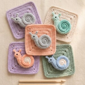 May include: A collection of five crocheted snail appliqu&eacute;s in various pastel colors, including lavender, peach, and mint green, each attached to a square base. The snails have detailed spiral shells and small antennae. Wooden knitting needles are at the bottom.