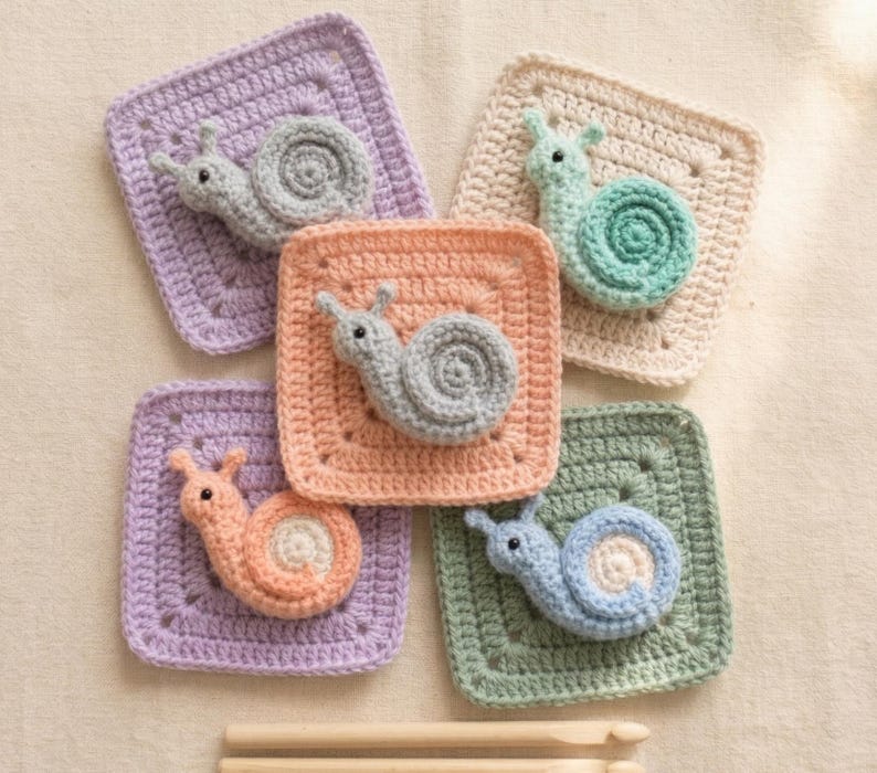 May include: A collection of five crocheted snail appliqu&eacute;s in various pastel colors, including lavender, peach, and mint green, each attached to a square base. The snails have detailed spiral shells and small antennae. Wooden knitting needles are at the bottom.