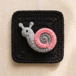 May include: A crocheted square with a black background features a gray snail with a pink and gray shell. The snail is centered on the square, with its body and shell details clearly visible. The square is made of yarn.