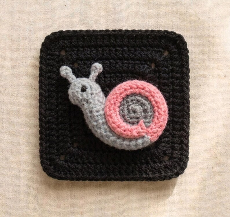 May include: A crocheted square with a black background features a gray snail with a pink and gray shell. The snail is centered on the square, with its body and shell details clearly visible. The square is made of yarn.