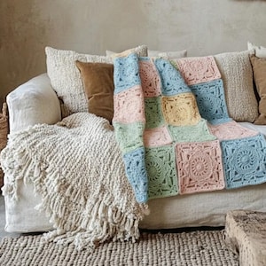 Könnte beinhalten: Eine gehäkelte Decke in Pastellfarben, bestehend aus quadratischen Feldern in Blau, Rosa, Grün und Gelb, drapiert über einem weißen Sofa. Das Sofa ist mit Kissen in neutralen Tönen ausgestattet. Ein Fransen-Wurf und ein gewebter Teppich sind ebenfalls zu sehen.