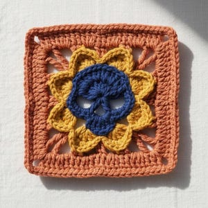 May include: Hand-crocheted square with a blue skull motif at the centre, encircled by yellow petals and a rust-coloured border. The textured piece is suitable for craft projects or decorative purposes.