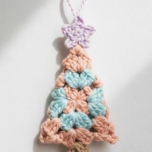 May include: A handmade crochet Christmas tree ornament. The tree is made of peach, light blue, and brown yarn, with a lavender star on top. The ornament has a loop for hanging. A festive, handcrafted decoration.