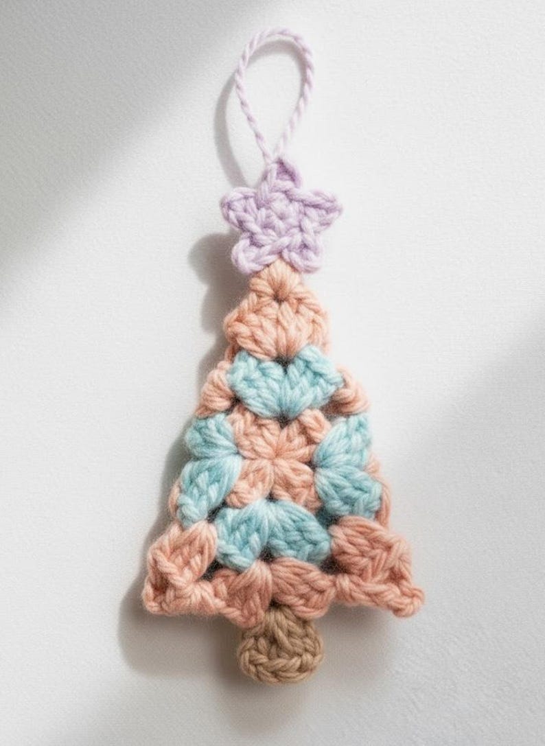 May include: A handmade crochet Christmas tree ornament. The tree is made of peach, light blue, and brown yarn, with a lavender star on top. The ornament has a loop for hanging. A festive, handcrafted decoration.