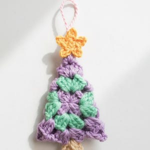 May include: A handmade crochet Christmas tree ornament. The tree is purple and green, topped with an orange star and a pink loop for hanging. The base of the tree is tan.