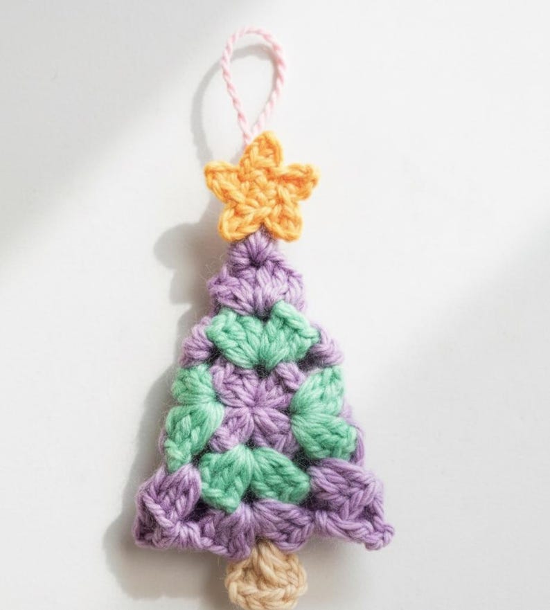 May include: A handmade crochet Christmas tree ornament. The tree is purple and green, topped with an orange star and a pink loop for hanging. The base of the tree is tan.