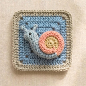 May include: A crocheted square featuring a light blue snail with a pink and tan shell. The snail is on a light blue background, framed by a gray border. The handmade item is likely part of a larger craft project.