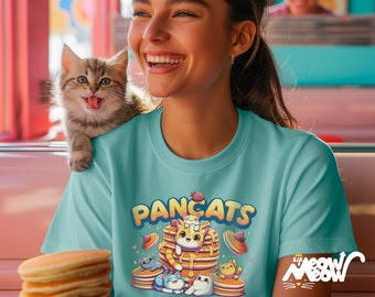 Könnte beinhalten: Ein türkisfarbenes T-Shirt mit einem Aufdruck von Katzen, die auf Pfannkuchen gestapelt sind. Der Text "Pancats" befindet sich über dem Aufdruck. Der Aufdruck enthält auch eine Erdbeere, Herzen und Sterne. Das T-Shirt wird von einer Person mit einem Kätzchen auf der Schulter getragen.