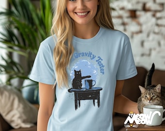 May include: A light blue t-shirt with a black cat graphic and the text "Gravity Tester". The cat is reaching for a cup of coffee on a table.