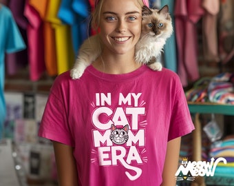 Peut inclure: Une femme portant un t-shirt rose avec le texte "In My Cat Mom Era" et un chat assis sur son épaule. Le chat est un Ragdoll avec des yeux bleus. La femme sourit et regarde la caméra.