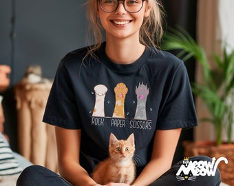 May include: A woman wearing a black t-shirt with a graphic of cat paws that says "Rock Paper Scissors". She is holding a small orange kitten.