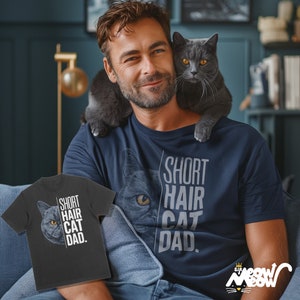 May include: A man wearing a navy blue t-shirt with the text "Short Hair Cat Dad." A gray cat is sitting on his shoulder. The man is also wearing a gray t-shirt with the same text and a cat image.